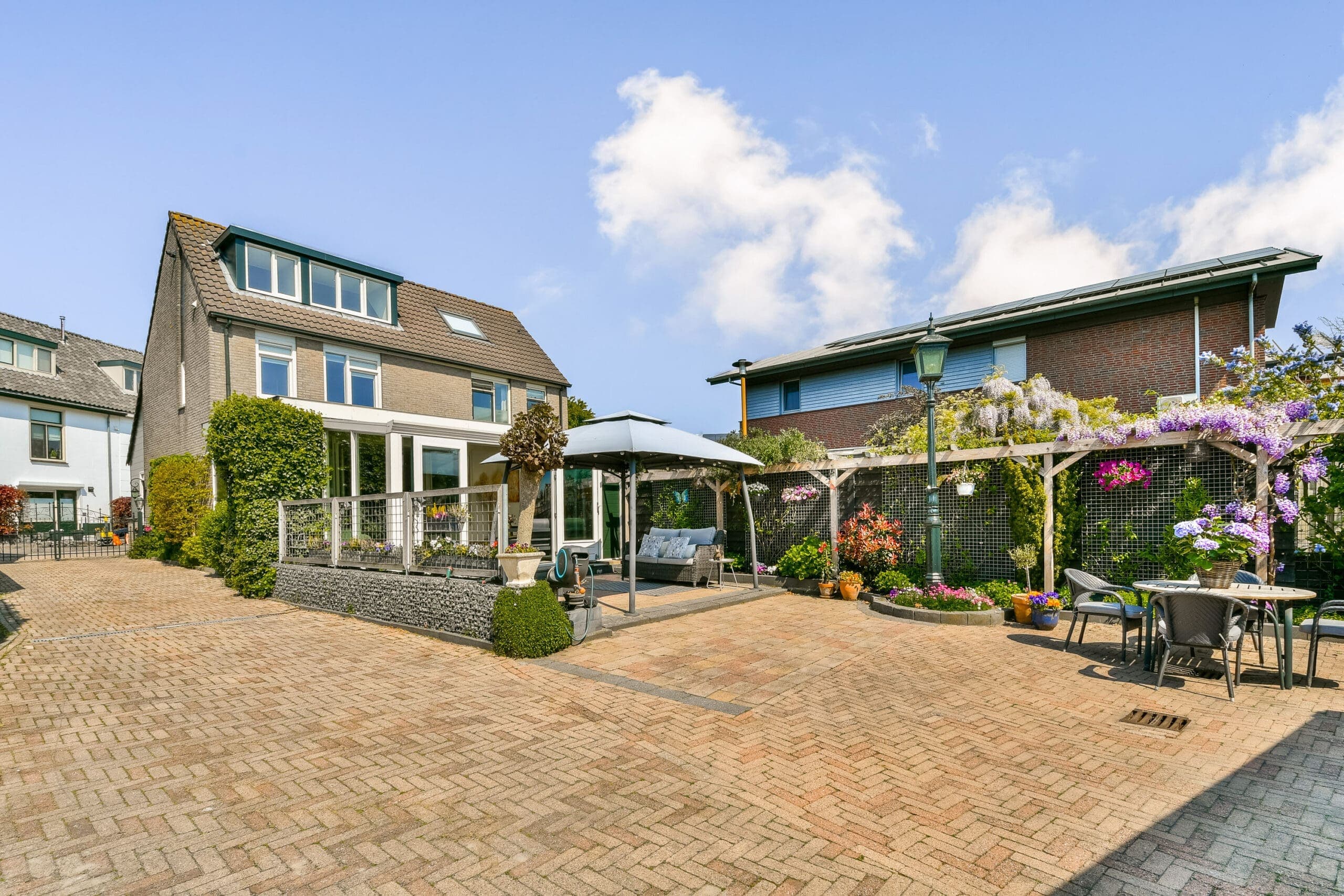 Woning met een terras onder een pergola met blauweregen, een loungehoek en een gemetselde tuin op een zonnige dag